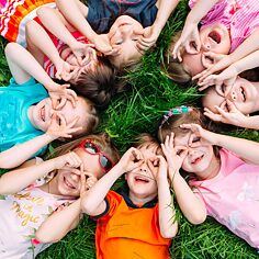 Eine Gruppe von Kindern liegt kreisförmig auf einer Wiese, sie halten die Hände über die Augen und grinsen in die Kamera