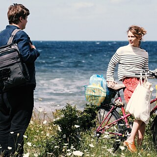 Filmszene aus Christian Petzolds Film "Roter Himmel": Nadja, gespielt von Paula Beer, steht mit einem Fahrrad an der Küste, im Hintergrund ist das Meer zu sehen. Sie lächelt Leon an, gespielt von Thomas Schubert, der mit dem Rücken zur Kamera steht und sich zu Nadja wendet.