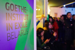 Purple-green roll-up with the lettering Goethe-Institut in Exile Belarus with audience in the background