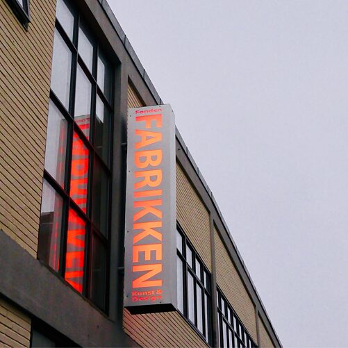 Auf dem Foto ist das Gebäude von FABRIKKEN for Kunst og Design von außen zu sehen, auf einem Neonschild steht "FABRIKKEN for Kunst og Design", die Schrift spiegelt sich in einem der Fenster.