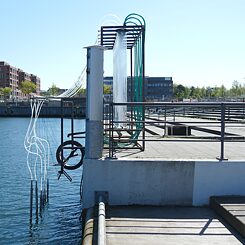 Seitenansicht einer Klangkunstinstallation im Hafen, bestehend aus sechs durchsichtigen Röhren, die über Schläuche mit dem Wasser verbunden und mit Salzwasser gefüllt sind. 
