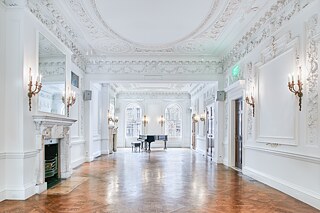 Photo of a brightly lit ballroom with ornate trim on the ceiling and walls. Lit candelabra sconces frame several entrances and mirrors above two white marble fireplaces. In the background near the windows, sits a grand piano and piano bench. 