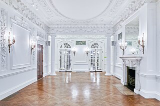 Photo of a brightly lit ballroom with ornate trim on the ceiling and walls. Lit candelabra sconces frame several entrances, a white marble fireplace, and two sets of mirrored french doors. 