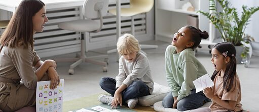 Junge Frau mit Plakat in der Hand sitzt drei Kindern gegenüber, die Aussprache der Buchstaben üben