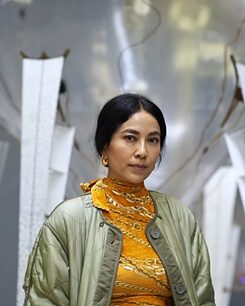 Anicka stands in front of a grey/silver artwork wearing a yellow roll neck and green satin jacket