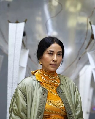 Anicka stands in front of a grey/silver artwork wearing a yellow roll neck and green satin jacket