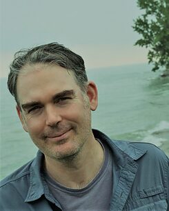 Keith steht vor einer malerischen Meereskulisse und trägt ein graues, aufgeknöpftes Hemd und ein T-Shirt