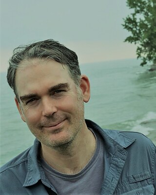 Keith steht vor einer malerischen Meereskulisse und trägt ein graues, aufgeknöpftes Hemd und ein T-Shirt