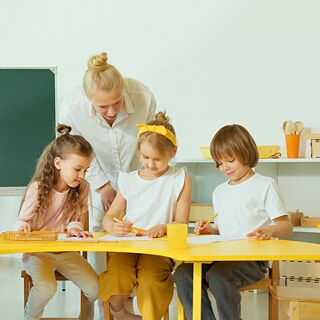 Schüler:innen der Grundschule sitzen an einem Tisch. Ihre Lehrerein steht daneben und schaut auf ihre Arbeiten