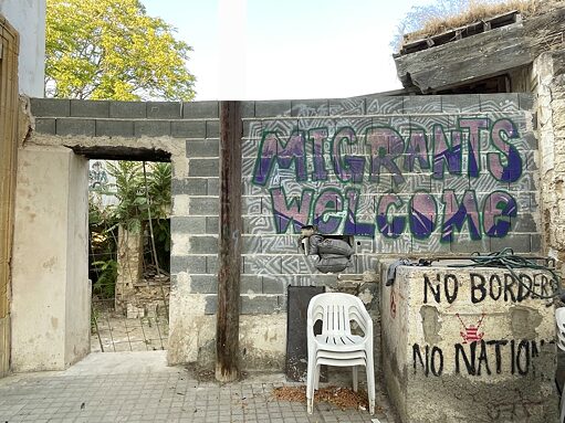 Eine Wand aus Ziegelsteinen mit den Schriften "Migrants welcome" und "No borders, no nations". Vor der Mauer sind weiße Plastikstühle gestapelt.