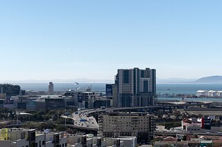 Der Nelson-Mandela-Boulevard schlängelt sich um die Hochhäuser der Stadt herum und endet an der Waterfront. Unten links im Bild ist eine relativ neue Siedlung zu sehen, in der Bewohner leben, die während der Apartheid aus dem District Six zwangsumgesiedelt wurden. Im Hintergrund ist die Table Bay zu sehen.