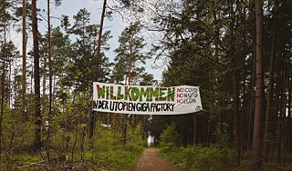 The photo shows a path in a pine forest. A banner is stretched between two trees. It states "Welcome to the Utopia Gigafactory. No Cops, no Nazis, no Elon"