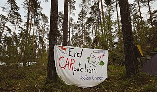 The photo shows a banner stretched between two pine trees. It states: "End CARpitalism, System Change"