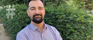 The picture shows Ervin Malakaj, a man in a chequered shirt with a beard, standing in front of a hedge. The logo of the Vorzeichen series can be seen at the top left of the picture.