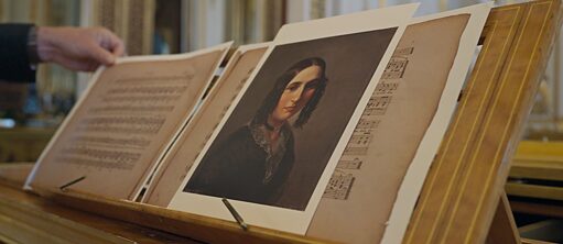 Foto von einem Musikständer auf einem Klavier mit einem Buch mit Noten, rechts ein Potraitbild von einer Frau mit einer Frisur aus dem 19. Jh.