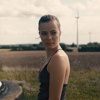 Eine junge Frau steht in einem Feld, im Hintergrund Windturbinen
