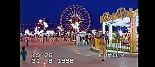 Auf dem Bild ist eine Szene von einem Jahrmarkt zu sehen, es ist dunkel, rechts ist ein Karussell zu sehen, im Hintergrund ein Riesenrad. Das Bild stammt aus einer Videoaufnahme, auf dem Bild sind das Datum 31.8.1998, die Uhrzeit 19:26 Uhr und "Familiy" angegeben. 