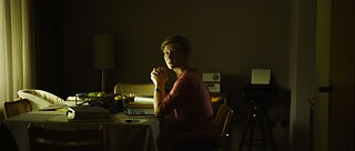 A woman sits at a table in front of a laptop in a dark room and looks at the camera