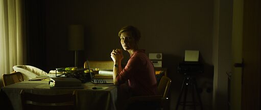 Eine Frau sitzt am Tisch vor einem Laptop in einem dunklen Zimmer und schaut an die Kamera