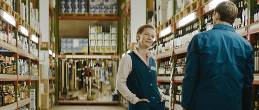 A man and a woman in work uniforms stand in a corridor of a store filled with products. They are facing each other as if in conversation.