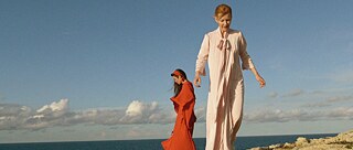 Δύο γυναίκες με μακριά φορέματα στέκονται σε βράχους. Στο βάθος διακρίνεται ένας συννεφιασμένος μπλε ουρανός και η θάλασσα. 
