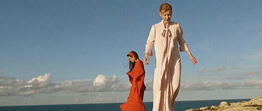 Two women in long dresses are standing on cliffs. A cloudy blue sky and the sea can be seen in the background. 