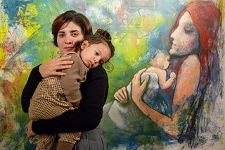Künstlerin steht mit einem Kind im Arm vor einer Malerei