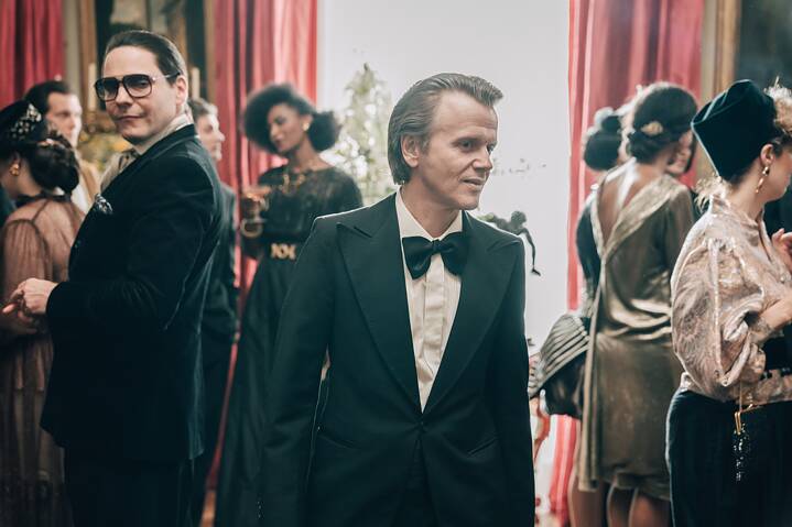 In einer Szene aus der Serie "Becoming Karl Lagerfeld" ist Pierre Bergé mittig im Bild im Rahmen einer Gesellschaftsrunde zu sehen, Karl Lagerfeld am Bildrand ihm hinterherblickend.