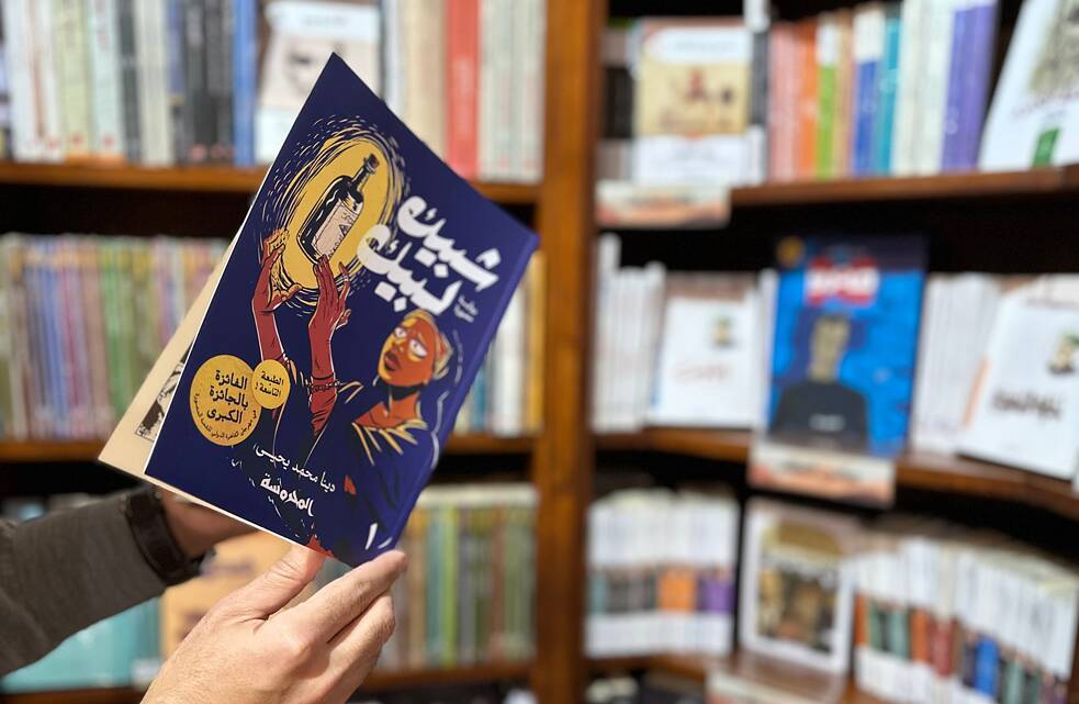 Une femme tient une bande dessinée. Le titre indique "Shubeik Lubeik" en arabe et le nom de l'auteur Deena Mohamed.