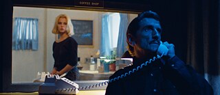 Man in the foreground on the phone, swathed in blue blackness, behind him a frame view into a coffee shop with a blonde waitress on the other end of the phone line 