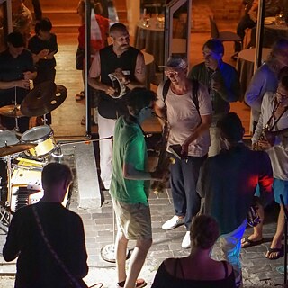 Das Bild zeigt eine Reihe von Jazzmusiker*innen, die bei der Summer Session gemeinsam jammen. Es ist dunkel im Raum und drumherum stehen viele Gäste.