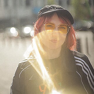 Die Pianistin und Komponistin Svetlana Marinchenko steht in einer Stadt vor einem Gebäude, das Sonnenlicht wird von der Kameralinse reflektiert. Ihre Haare sind rotgefärbt und schauen unter einer schwarzen Kappe hervor, sie trägt eine gelbe Sonnenbrille.