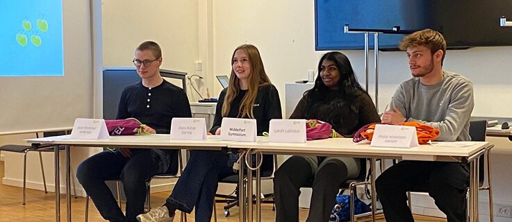 4 elever sidder ved et bord med navneskilte foran sig. De har sportstasker foran sig, som de har vundet i en debatkonkurrence.