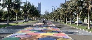 A street, the corniche walk of Jeddah, in Saudi Arabia with a pattern of colours. Two people are standing in the background talking to each other. The street is surrounded by palm trees. 
