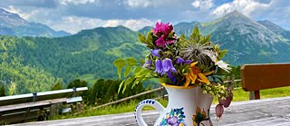 Ein alpenländischer Tonkrug mit einem bunten Blumenstrauß steht auf einem Tisch, im Hintergrund ist eine Alpenlandschaft zu sehen.