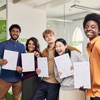 Das Bild zeigt eine Gruppe von fünf jungen Erwachsenen, die glücklich und stolz in die Kamera lächeln. Jeder von ihnen hält ein Goethe-Zertifikat B2 hoch. Sie stehen in einem modernen Raum mit großen Fenstern und strahlen Freude und Zufriedenheit aus. Ihre Kleidung ist bunt und unterschiedlich, was die Vielfalt der Gruppe betont.