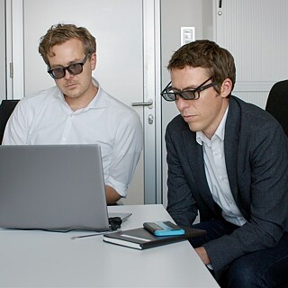 Two men wearing two pairs of glasses look at a screen in an office