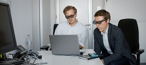 Two men wearing two pairs of glasses look at a screen in an office