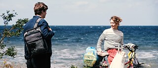 A scene from the movie "Afire": Leon, played by Thomas Schubert, is standing on the left of the picture, carrying a black rucksack on his right shoulder and looking at Nadja. Nadja, played by Paula Beer, is standing on the right-hand edge of the picture with a bicycle loaded with shopping bags. They look at each other. Both are standing by the dune vegetation zone, with the Baltic Sea in the background. 