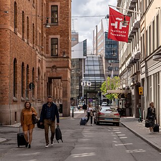 Eine Straße umringt von Häusern. Dort laufen mehrere Menschen, E-Roller stehen auf dem Bürgersteig. An einer Häuserwand hängen die Fahne des Goethe-Instituts.