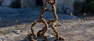 Abstract sculpture made of rope-like material, built on sand, with a kind of stone dwelling visible in the background. 