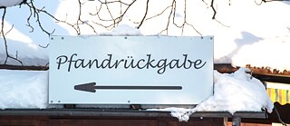 Ein Schild mit dem Wort „Pfandrückgabe" und einem Pfeil nach links, das mit Schnee bedeckt ist und die Richtung für die Rückgabe von Flaschen oder Dosen anzeigt. Im Hintergrund sind Äste und eine schneebedeckte Landschaft zu sehen.