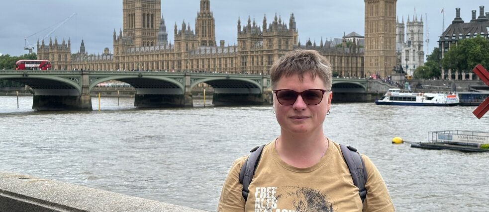 A woman wearing a beige t-shirt and sunglasses stands in front of the River Thames