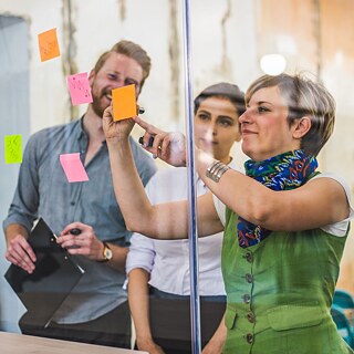  A woman points at a sticky note on a glass wall, highlighting a key message in a professional development seminar.