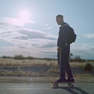 A white man rides a skateboard along a sunny country road in a wide open landscape.