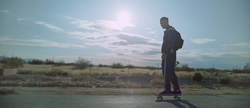 A white man rides a skateboard along a sunny country road in a wide open landscape.