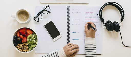 Eine Person schreibt in ein Notizblock. Auf dem Schreibtisch befindet sich eine Brille, ein Kopfhörer, ein Handy, ein Laptop sowie eine Tasse Café und eine Müsli-Schale mit Obst und Nüsse. Aufnahme von oben.