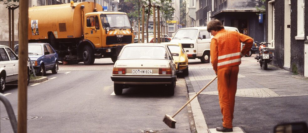 Auf dem Bild ist ein altes Dia zu sehen. Man sieht die Müllabfuhr in Dortmund. Im Vordergrund steht eine orange-gekleidete Person, die eine Straße kehrt. Im Hintergrund biegt ein Müllwagen um die Ecke.