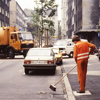 Auf dem Bild ist ein altes Dia zu sehen. Man sieht die Müllabfuhr in Dortmund. Im Vordergrund steht eine orange-gekleidete Person, die eine Straße kehrt. Im Hintergrund biegt ein Müllwagen um die Ecke.