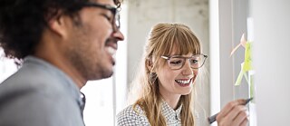 Zwei Kolleg*innen stehen nebeneinander im Büro vor einer Pinnwand. © © Getty Images Teamwork